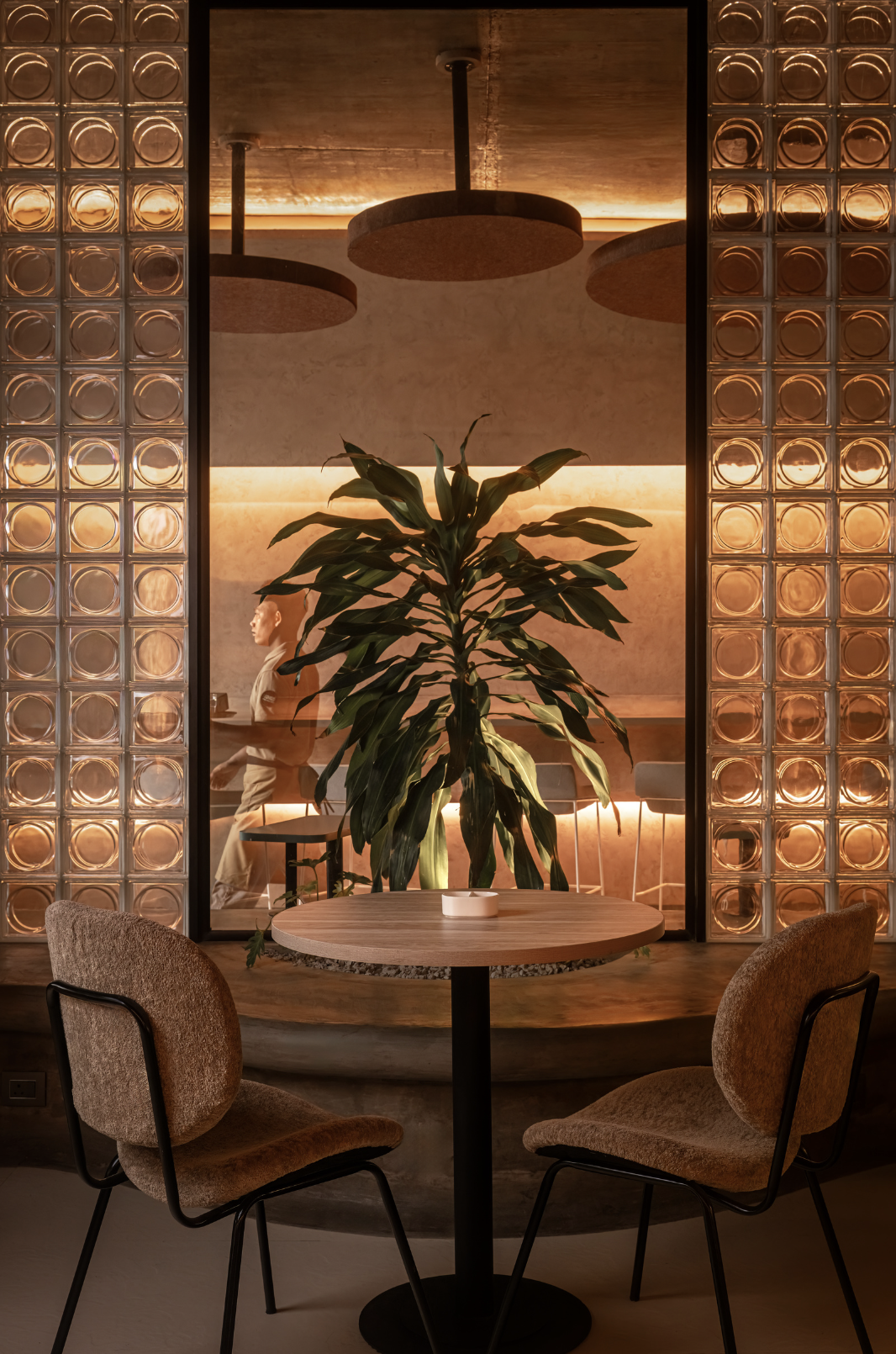 Warm cafe interior framed by glowing glass block columns, bouclé chairs, and lush greenery