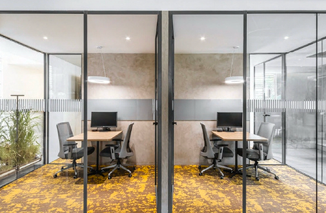 Twin glass meeting pods with wood desks, monitors, and patterned yellow carpet